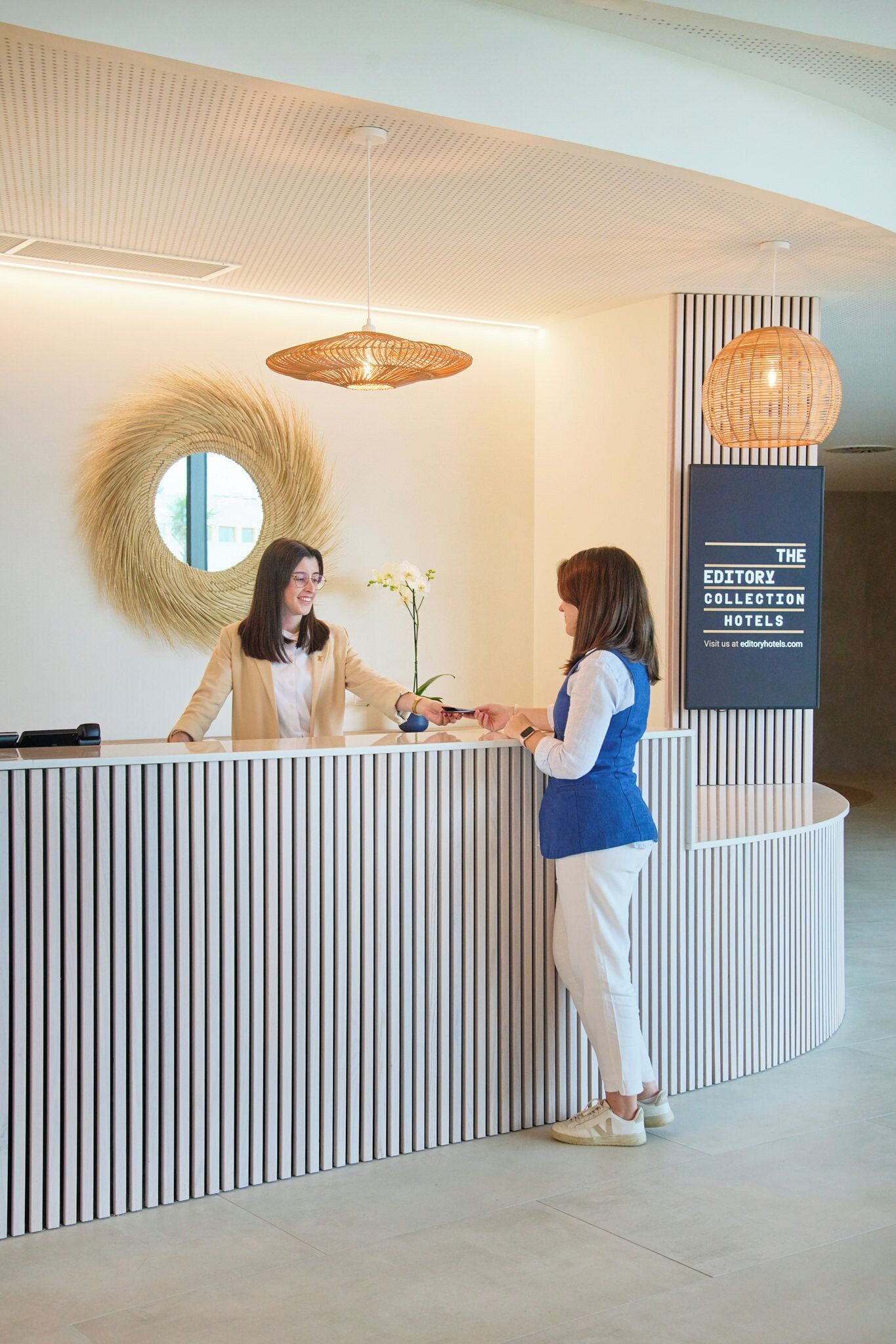 A reception desk with a modern design, featuring a circular mirror and a hanging light fixture. A signboard for 'The Editory Collection Hotels' is visible.