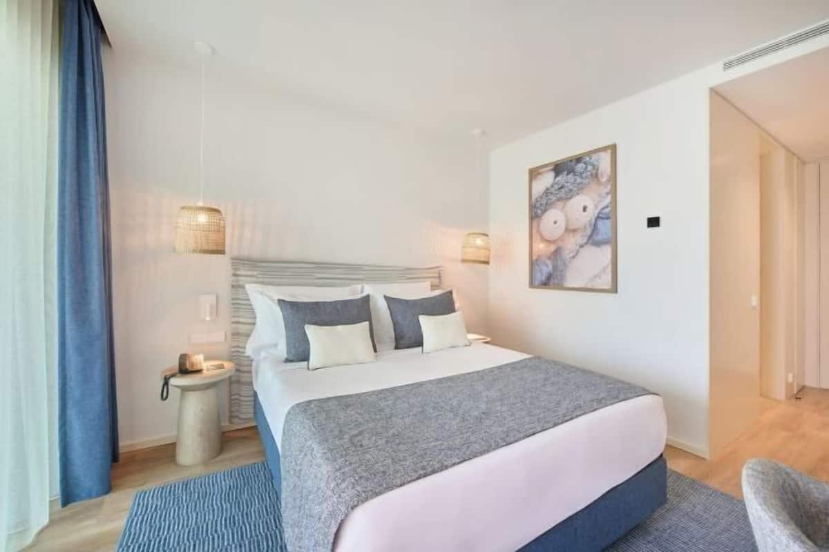 Ein modernes Hotelzimmer mit einem großen Bett, Nachttischlampen, einem kleinen Nachttisch und einem Gemälde an der Wand.