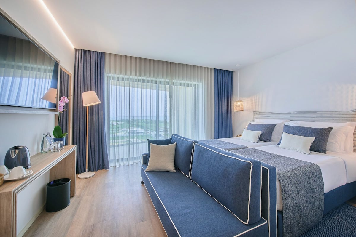 Ein modernes Hotelzimmer mit einem großen Bett, einer Couch, einem Schreibtisch mit Lampe und Blick ins Freie.