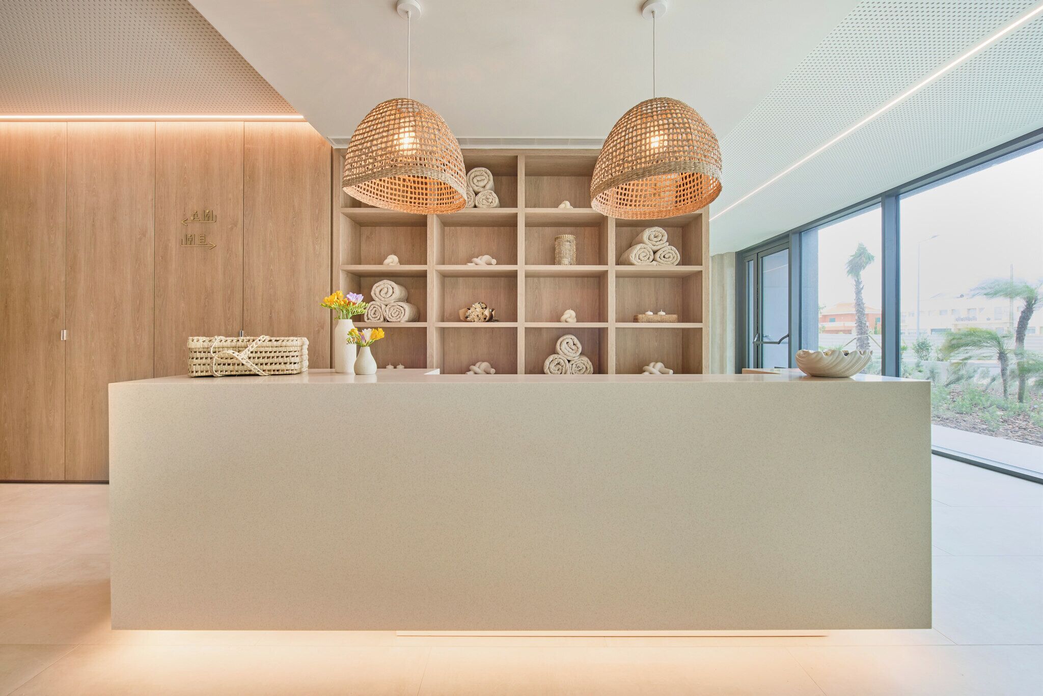 A reception desk with a neutral color scheme, two woven pendant lights, and a view of the outdoors through large windows.