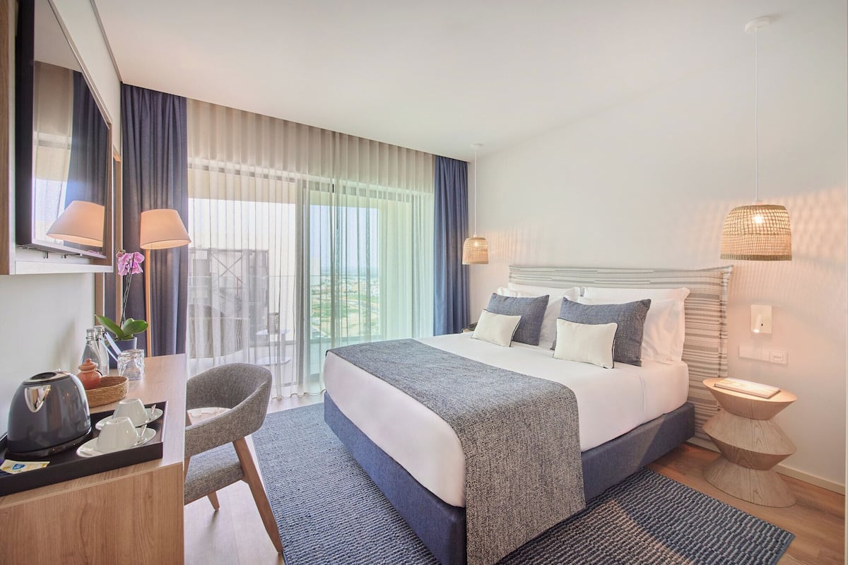 Ein modernes Hotelzimmer mit einem großen Bett, einem Schreibtisch mit Kaffeemaschine, einem Sessel und Blick ins Freie durch große Fenster.