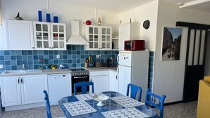 A compact kitchen with white cabinets, a stove, and a refrigerator. There is a dining table with blue chairs and a patterned tablecloth. - Charming ground-floor apartment 150m from the beach  (Saint-Georges-de-Didonne)