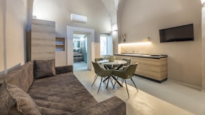 Een moderne woonkamer met een bank, eettafel en stoelen. Er is een keukenhoek met een televisie en een raam.