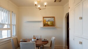 Een eetkamer met een ronde tafel, vier stoelen, een kroonluchter, een schilderij aan de muur en een raam met jaloezieën.