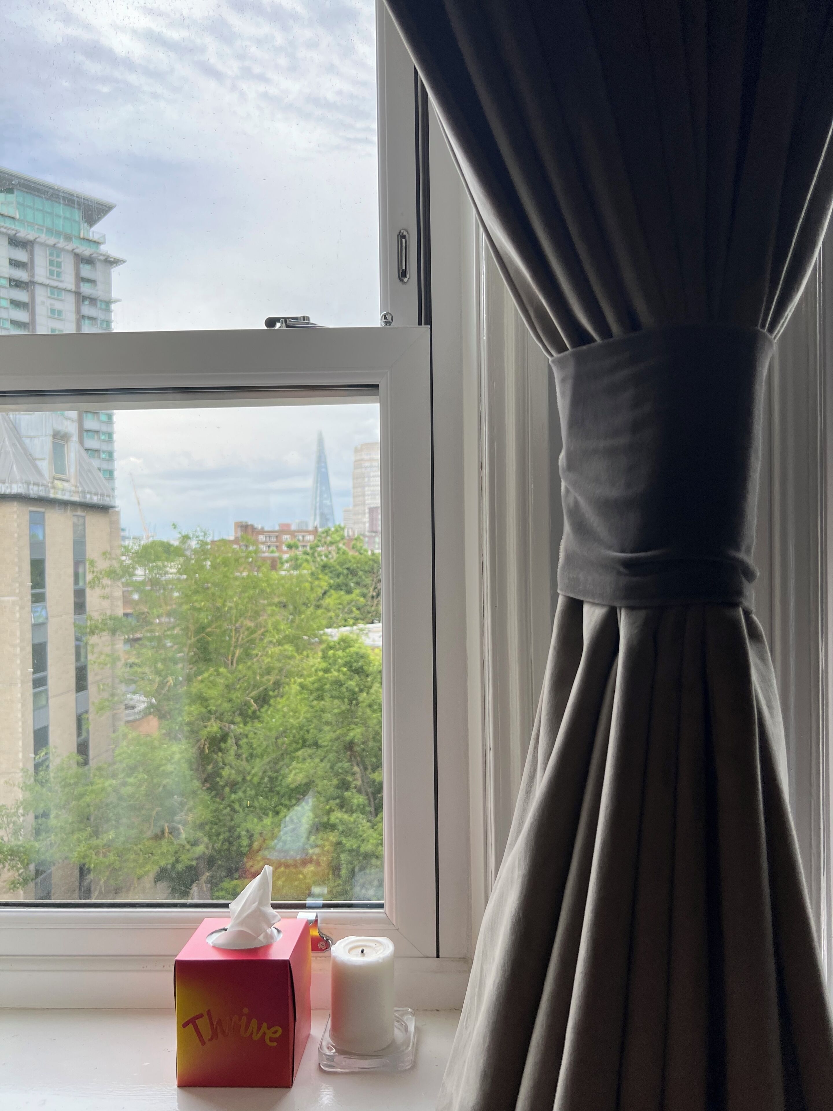 A window with a view of buildings and trees, a tissue box, and a candle on the windowsill.