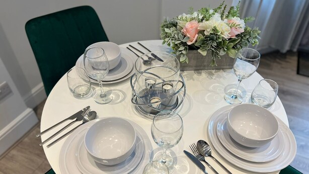 A round dining table set with white plates, bowls, and clear glassware, accompanied by a floral centerpiece and green upholstered chairs.