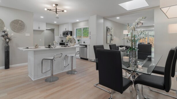 Una cocina moderna con armarios blancos, una isla central con taburetes y un comedor con mesa de vidrio y sillas negras.