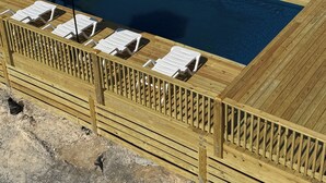 Una piscina rectangular con cubierta de madera, rodeada de sillas reclinables blancas y dos sombrillas azules.
