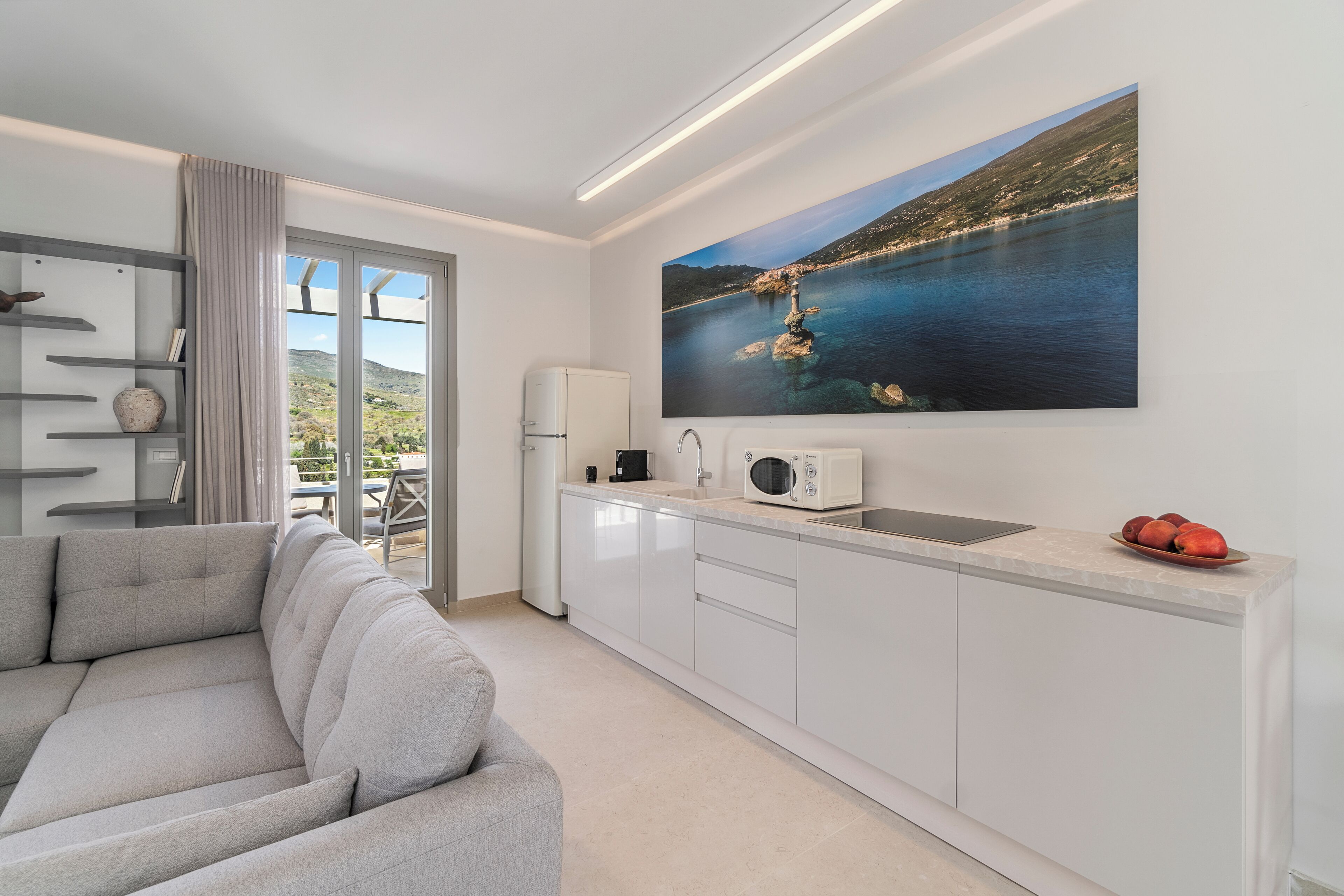 A modern kitchen with a large window, a grey sofa, and a painting of a coastal landscape.