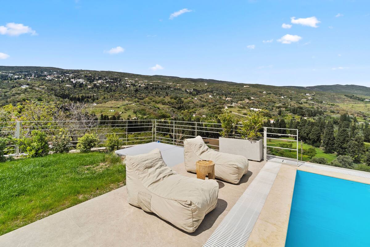 Uma área à beira da piscina com um sofá bege e almofadas, um banquinho de madeira e vista para uma paisagem montanhosa.