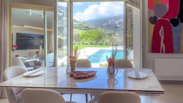 Un comedor moderno con una mesa de vidrio, sillas blancas y vistas a una piscina y vegetación.