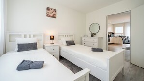A modern hotel room with two single beds, a bedside table with a lamp, a small dresser, and a large mirror.