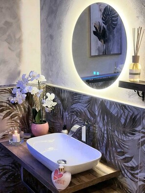 Ein Badezimmer im modernen Stil, mit einem großen, ovalen Waschbecken, einem Spiegel mit rundem Rahmen und einer Zierpflanze.