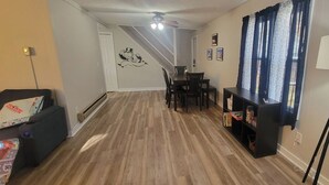 Een woonkamer met een houten vloer, een zwarte bank, een eettafel met stoelen, een zwarte boekenkast en een plafondventilator.