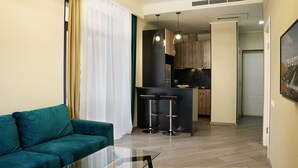 A modern living room with a green sofa, a glass coffee table, and a kitchen area with wooden cabinets and a black island. - 1BR Getaway w/ Pool & Sauna Access (Tsaghkadzor)