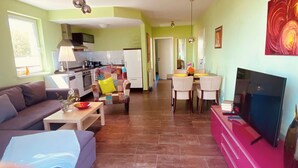 A living room with a sofa, a coffee table, and a TV. There is a kitchen in the background with a dining area. - House directly on the beach, behind the dune, sea view (Breege)