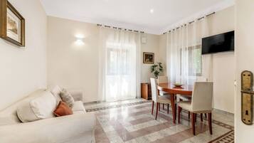A living room with a sofa, a dining table with chairs, a TV mounted on the wall, and a patterned floor.