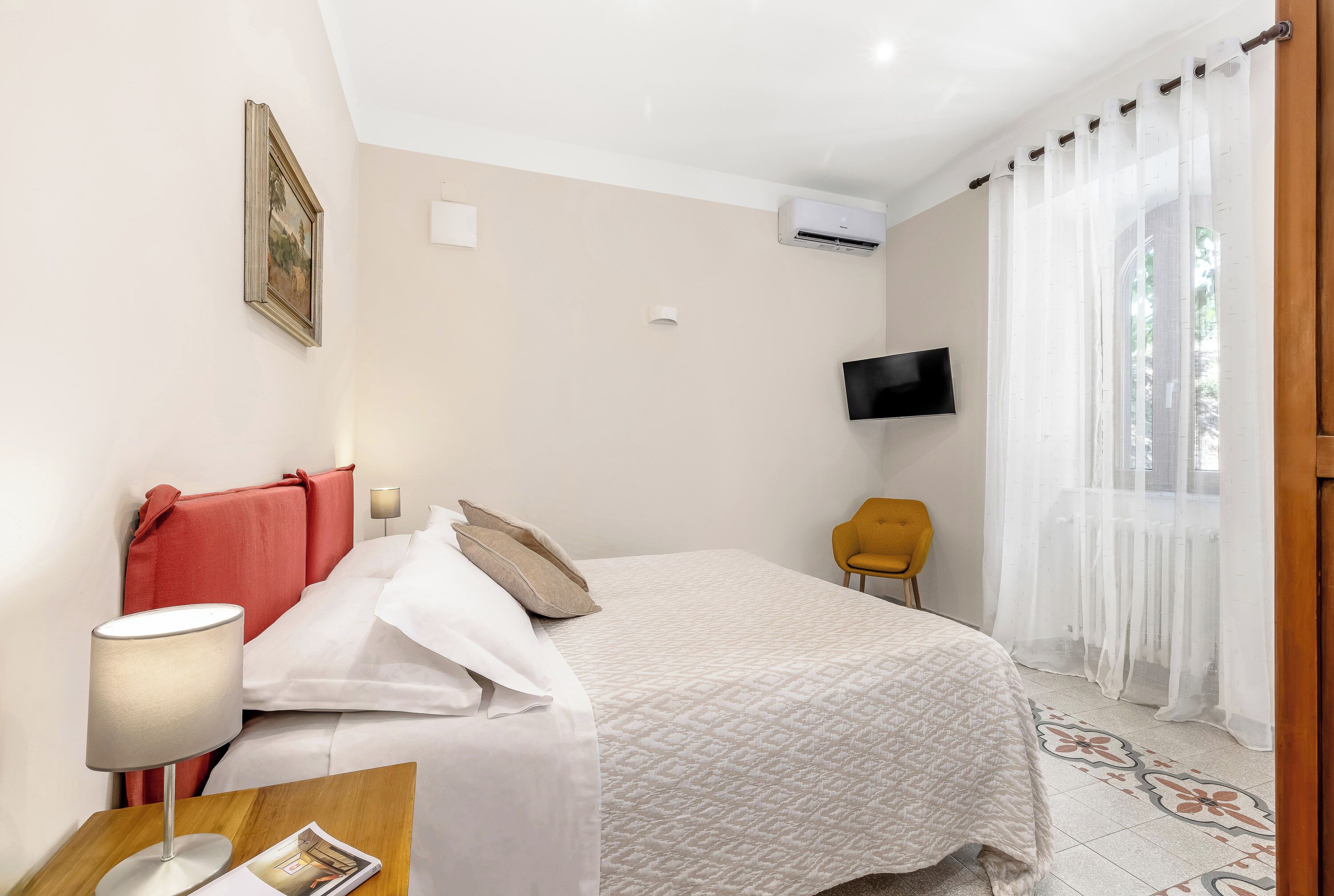 A hotel room with a bed, a TV, an air conditioning unit, a chair, and a lamp.