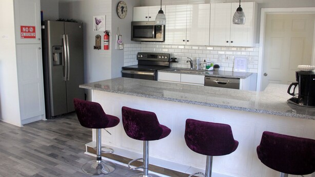 A modern kitchen with white cabinets, stainless steel appliances, and a marble countertop. There are three purple bar stools and a stainless steel refrigerator. - Charming 2-bedroom apartment with shared Pool, frontyard WiFi, AC (St. Petersburg)