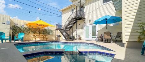 Un patio trasero con piscina, una escalera que lleva a un balcón en un segundo piso, muebles de exterior y un área cercada con plantas en macetas.