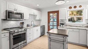 A modern kitchen with white cabinetry, stainless steel appliances, a large island, and a view through a glass door. - New Golf Cart-Private Hot Tub-Walk to Beach-Charming Bungalow 4BR! (Santa Rosa Beach)