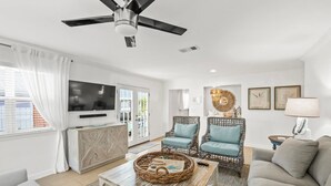 A modern living room with a ceiling fan, wicker coffee table, two blue armchairs, a grey sofa, and a TV on the wall. - New Golf Cart-Private Hot Tub-Walk to Beach-Charming Bungalow 4BR! (Santa Rosa Beach)