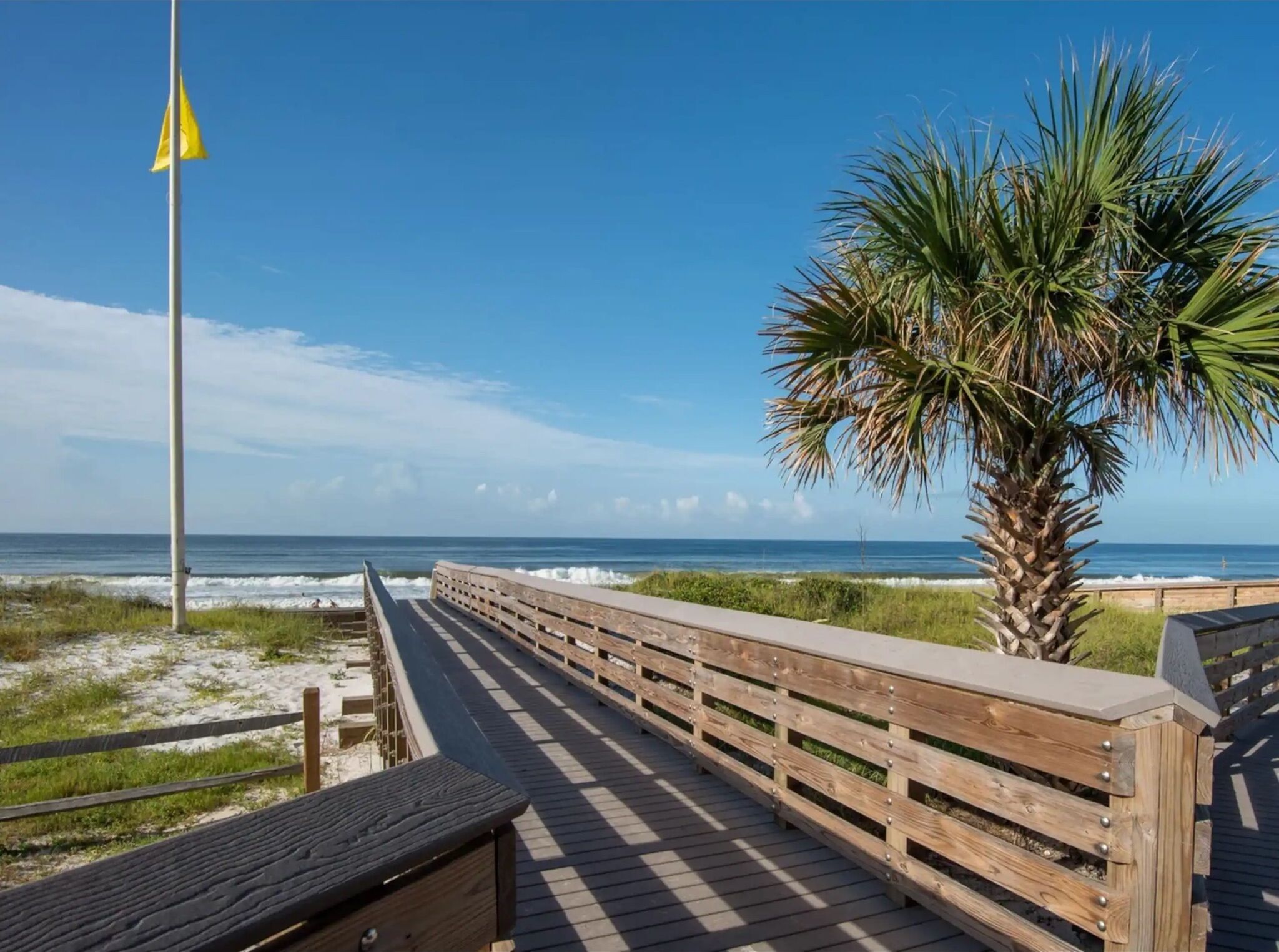 A wooden boardwalk leading to the beach with a palm tree and a flagpole.