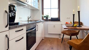 Een moderne keuken met witte kasten, een ingebouwde oven en een koffieapparaat. Er is een raam met uitzicht naar buiten, een tafel met een schaal fruit en twee kaarsen op tafel.