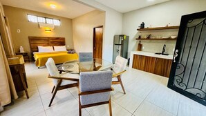 Een moderne hotelkamer met een bed, een zithoek met een glazen tafel en een keukenhoek met een koelkast en planken.