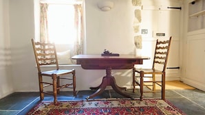 A dining area with a wooden table and two chairs, a patterned rug, a window with floral curtains, and a door with a coat rack. - Church Cottage (Exeter)