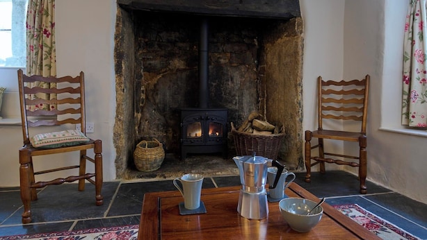 A cozy room with a stone fireplace, two wooden chairs, a table with a coffee pot, cups, and a bowl. - Church Cottage (Exeter)