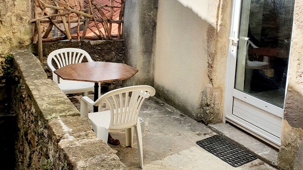 A stone-walled outdoor area with a table and chairs, a door with a glass panel, and a lantern. - Fully renovated 2 to 3 person gîte (Saint-Rome-de-Tarn)