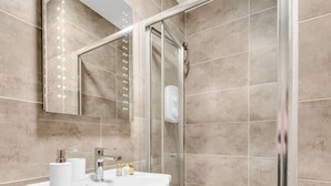 Une salle de bain moderne avec une douche à parois de verre, un lavabo blanc agrémenté d’articles de toilette et un grand miroir avec éclairage.
