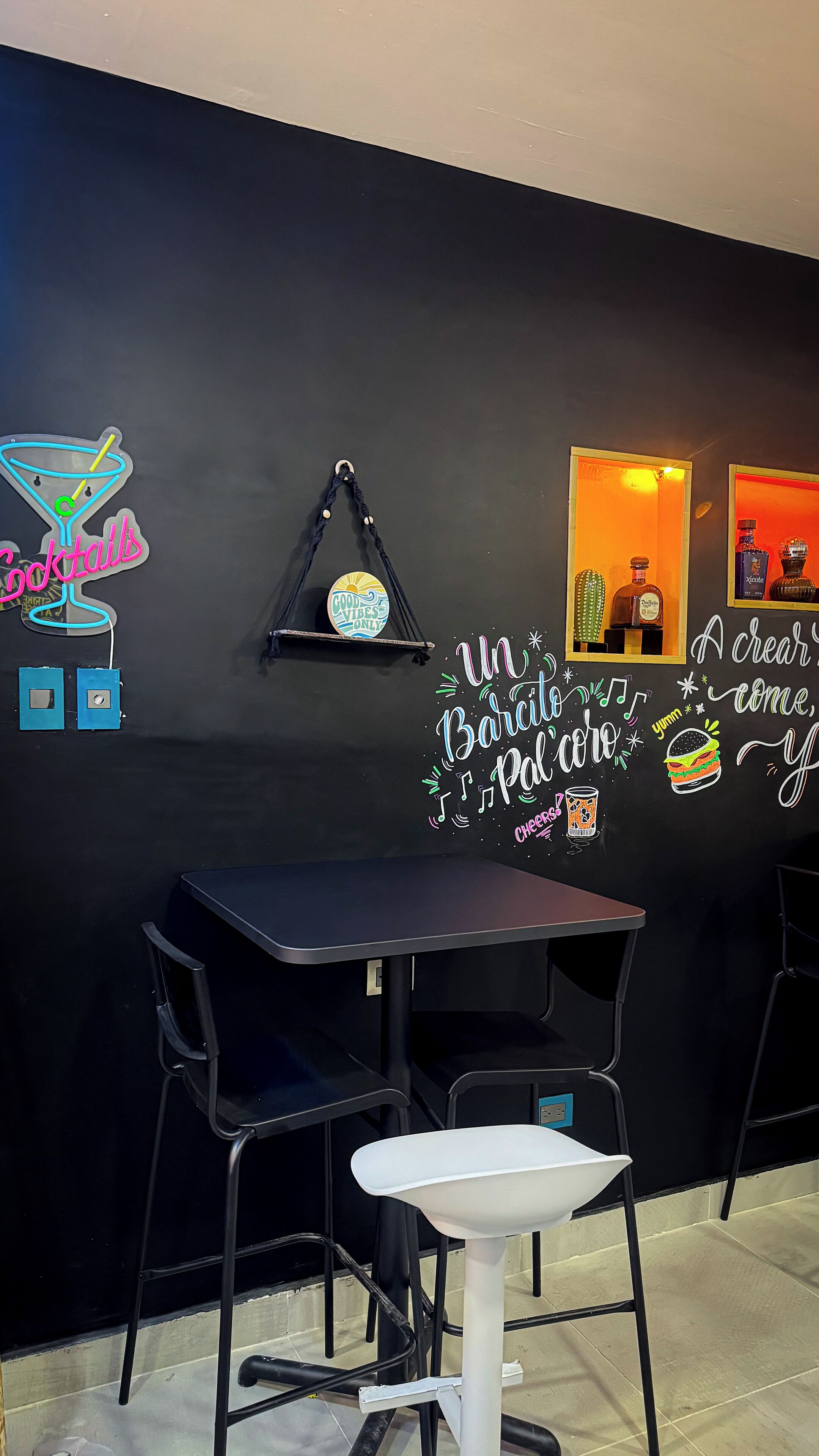 Uma área de bar com parede preta, luminosos de neon, um banquinho e uma mesa.