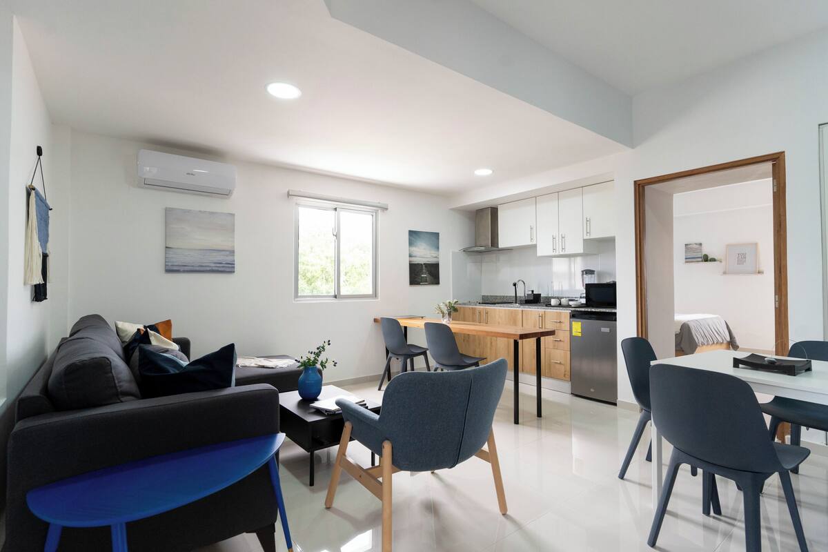 Una sala moderna con sofá, sillas y una mesa de comedor. Hay una zona de cocina con armarios y electrodomésticos.