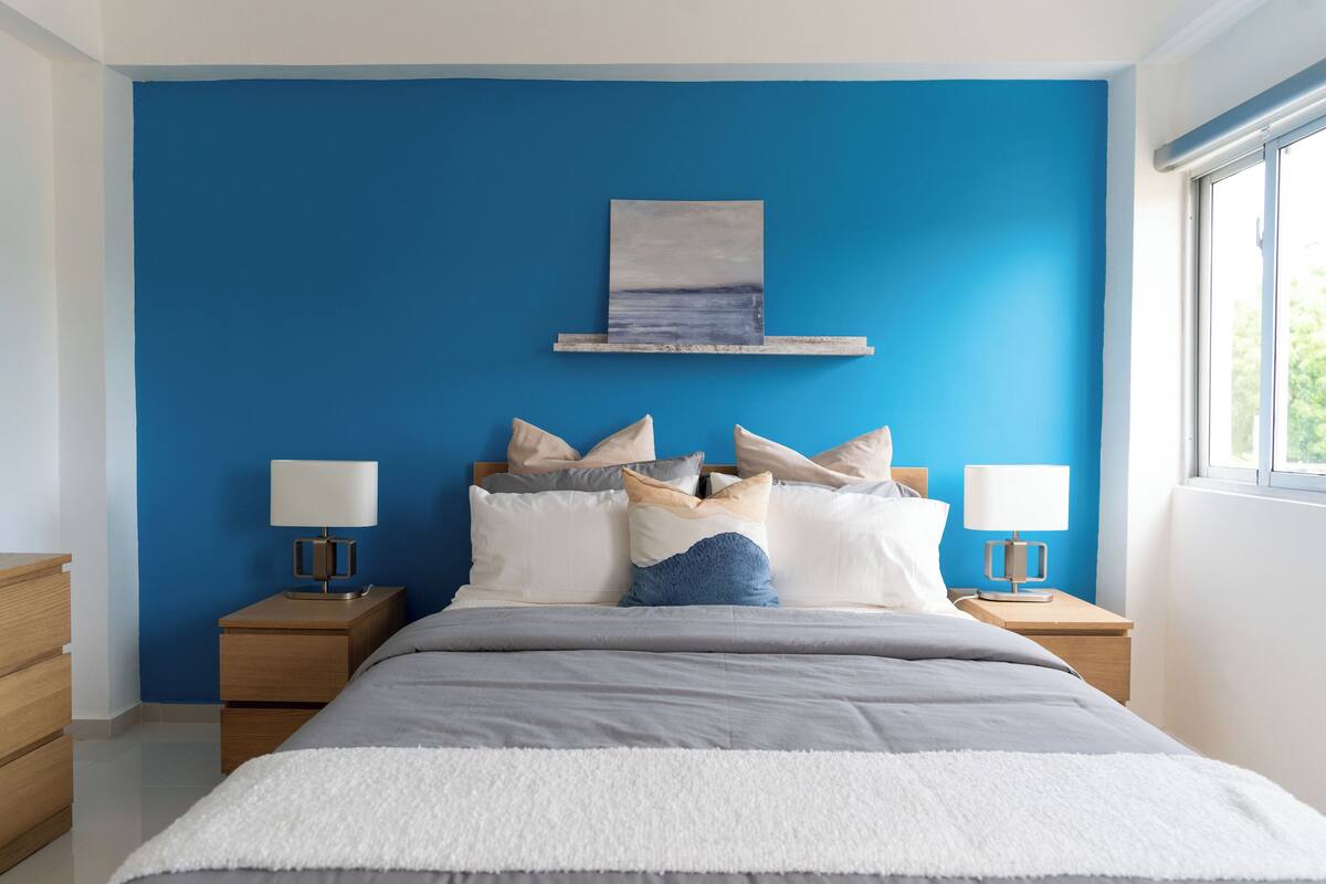 Un dormitorio con una pared azul destacada, una cama con muchas almohadas, dos mesitas de noche con lámparas y un cuadro sobre la cama.