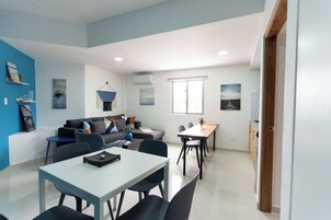 Una sala de estar moderna con un sofá, mesa de comedor y sillas. Hay estantes en la pared con objetos decorativos.