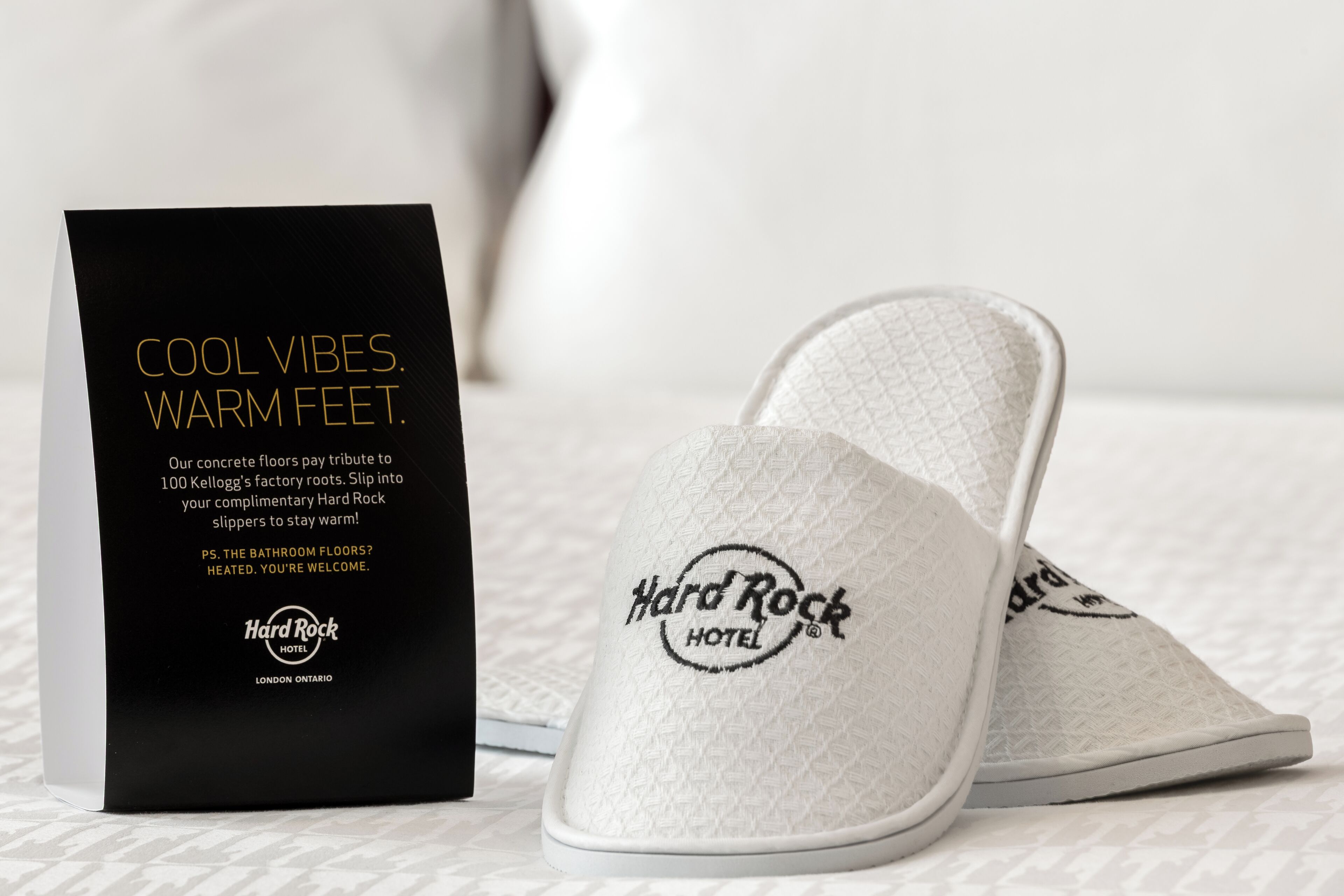 Ein Paar weiße, lockere Hausschuhe mit einer strukturierten Oberfläche und dem Logo des Hard Rock Hotels, neben einer schwarzen Box mit Werbung für die Hausschuhe.