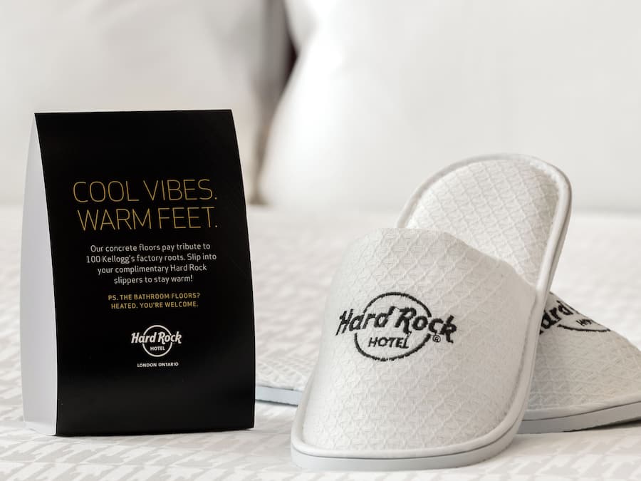 Ein Paar weiße, lockere Hausschuhe mit einer strukturierten Oberfläche und dem Logo des Hard Rock Hotels, neben einer schwarzen Box mit Werbung für die Hausschuhe.