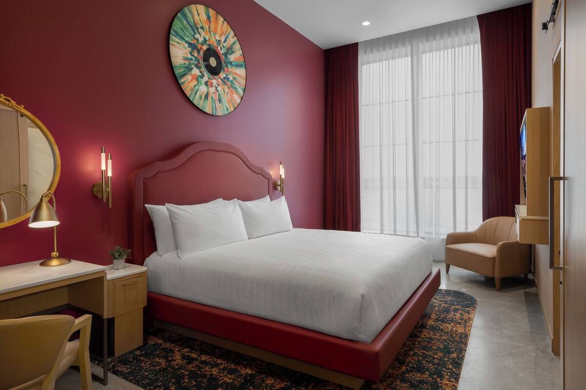 Une chambre d’hôtel avec un grand lit, un bureau avec une lampe, une chaise et une grande fenêtre avec des rideaux.