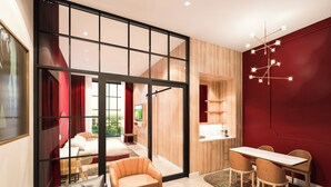 Una habitación de hotel moderna con un gran ventanal corredera que da a un balcón, un sofá, una mesa de centro, una zona de comedor con mesa y sillas, y una cocina integrada.