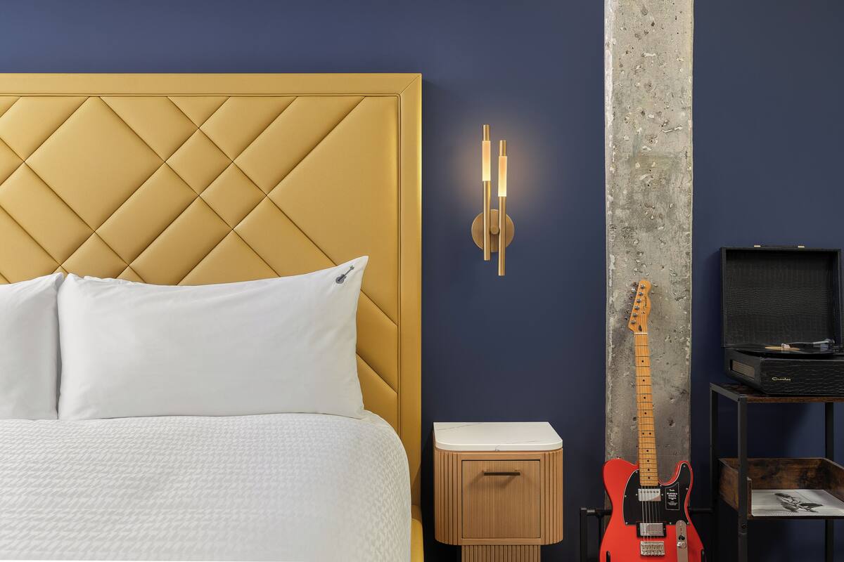 Une chambre à coucher avec une tête de lit capitonnée, une table de chevet, une guitare et un téléviseur posé sur un meuble.