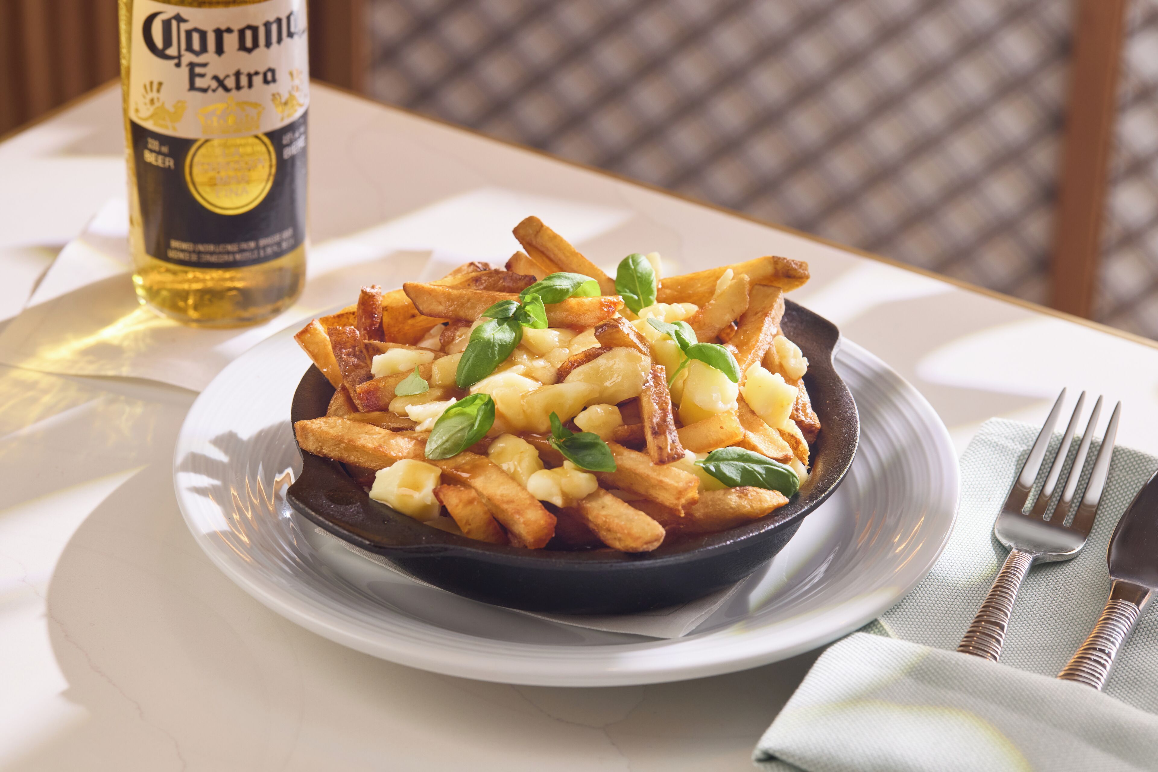 Un plato de papas fritas con queso y albahaca, una botella de cerveza Corona Extra, y un tenedor y cuchillo sobre una mesa.