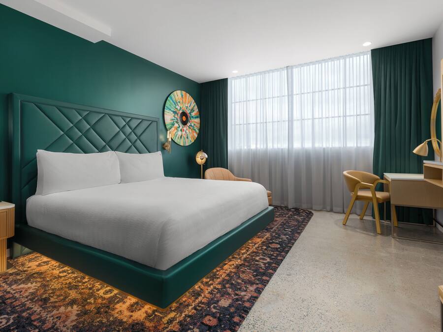 Ett modernt hotellrum med en stor säng, ett skrivbord, en stol och ett stort fönster med gardiner.