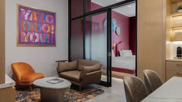 A modern hotel room with a sofa, armchair, dining table, and a large window.