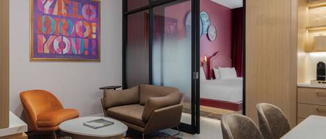 A modern hotel room with a sofa, armchair, dining table, and a large window.