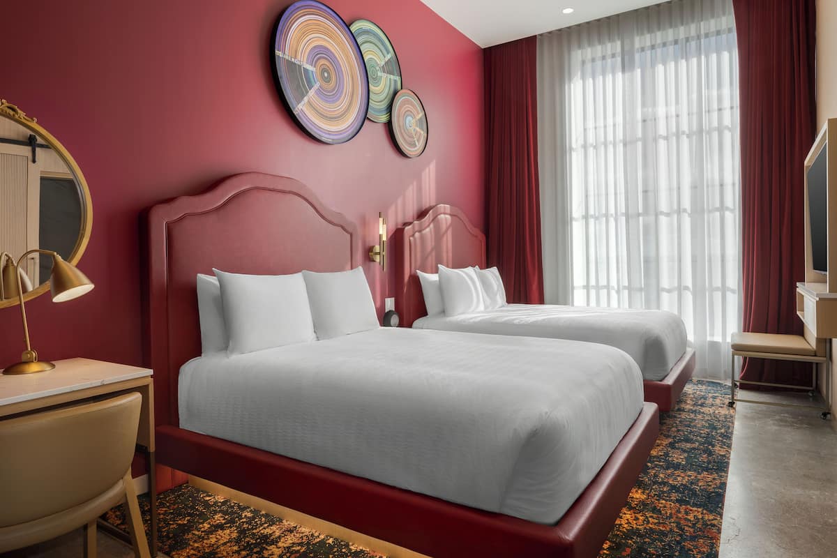 Une chambre d’hôtel avec deux lits, un bureau, une chaise, un miroir et une grande fenêtre avec des rideaux.