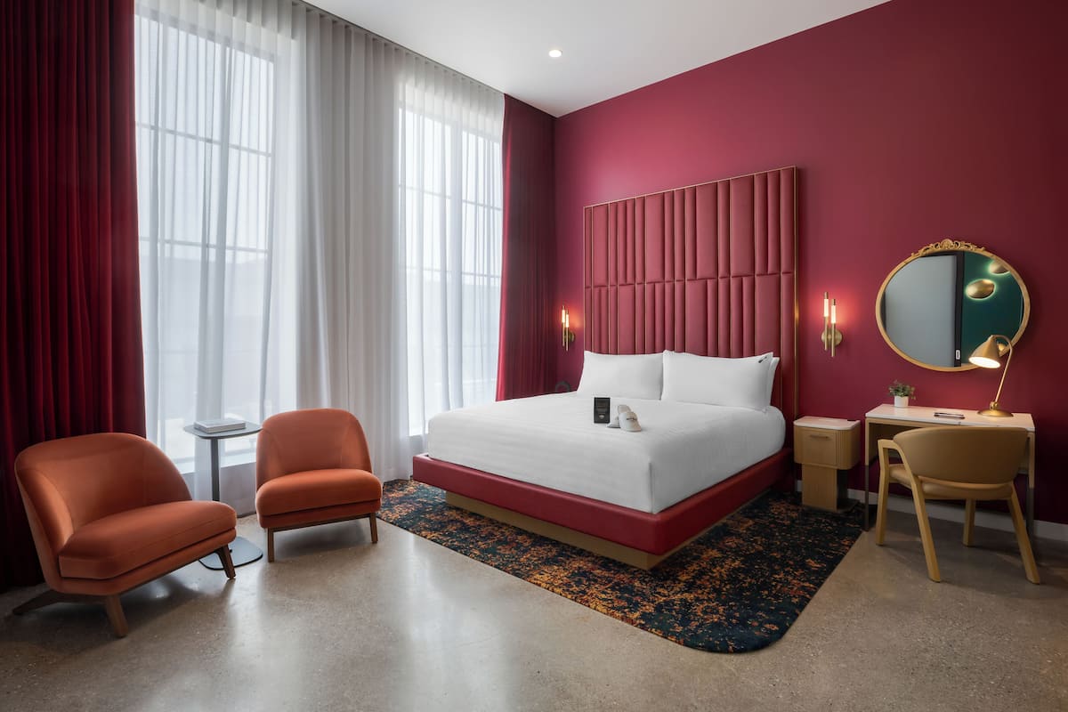 Une chambre d’hôtel avec un grand lit, deux fauteuils, un bureau avec une chaise, un miroir rond et une petite table.