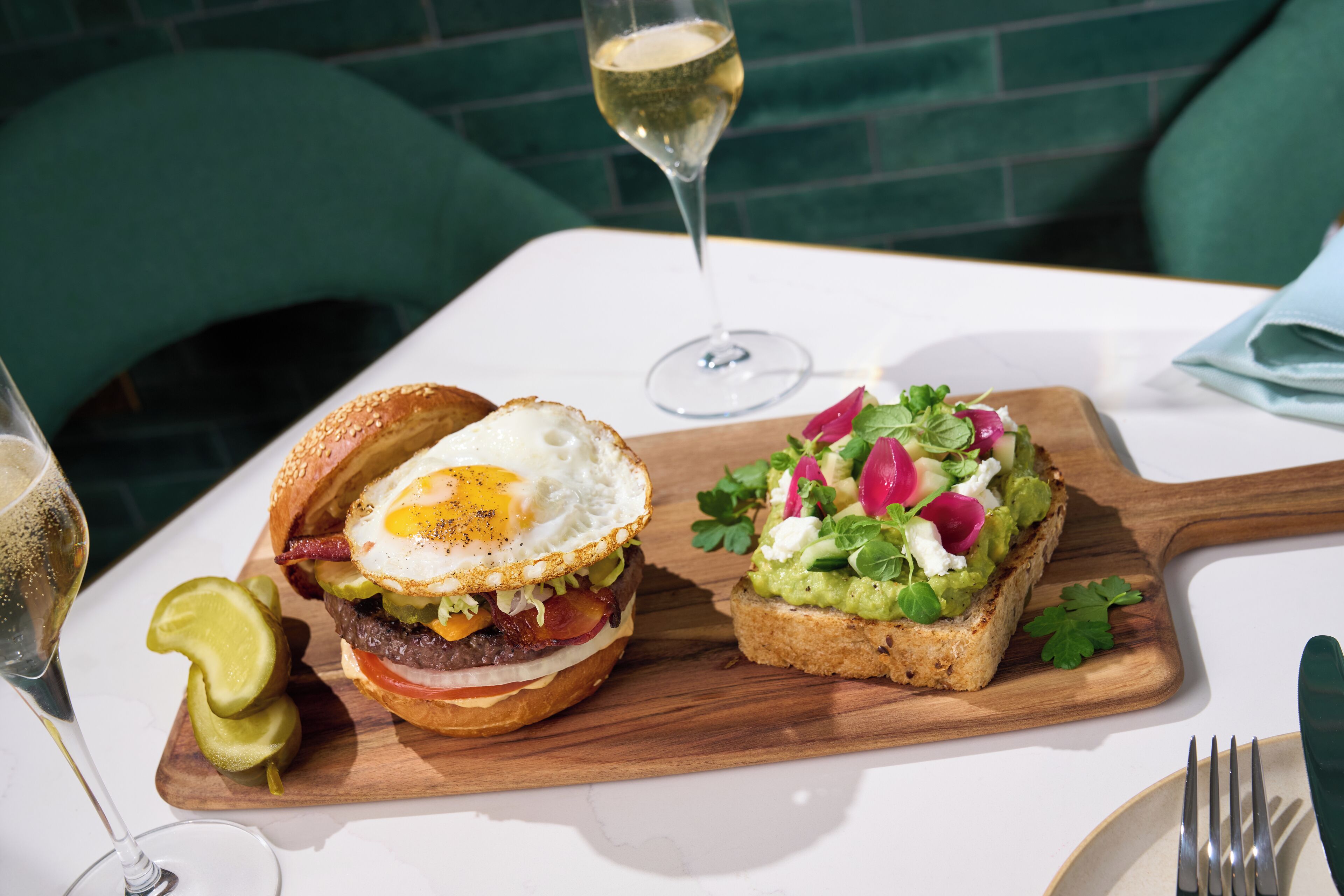 A meal with a burger and a sandwich on a wooden board, a glass of wine, and a fork on a table.