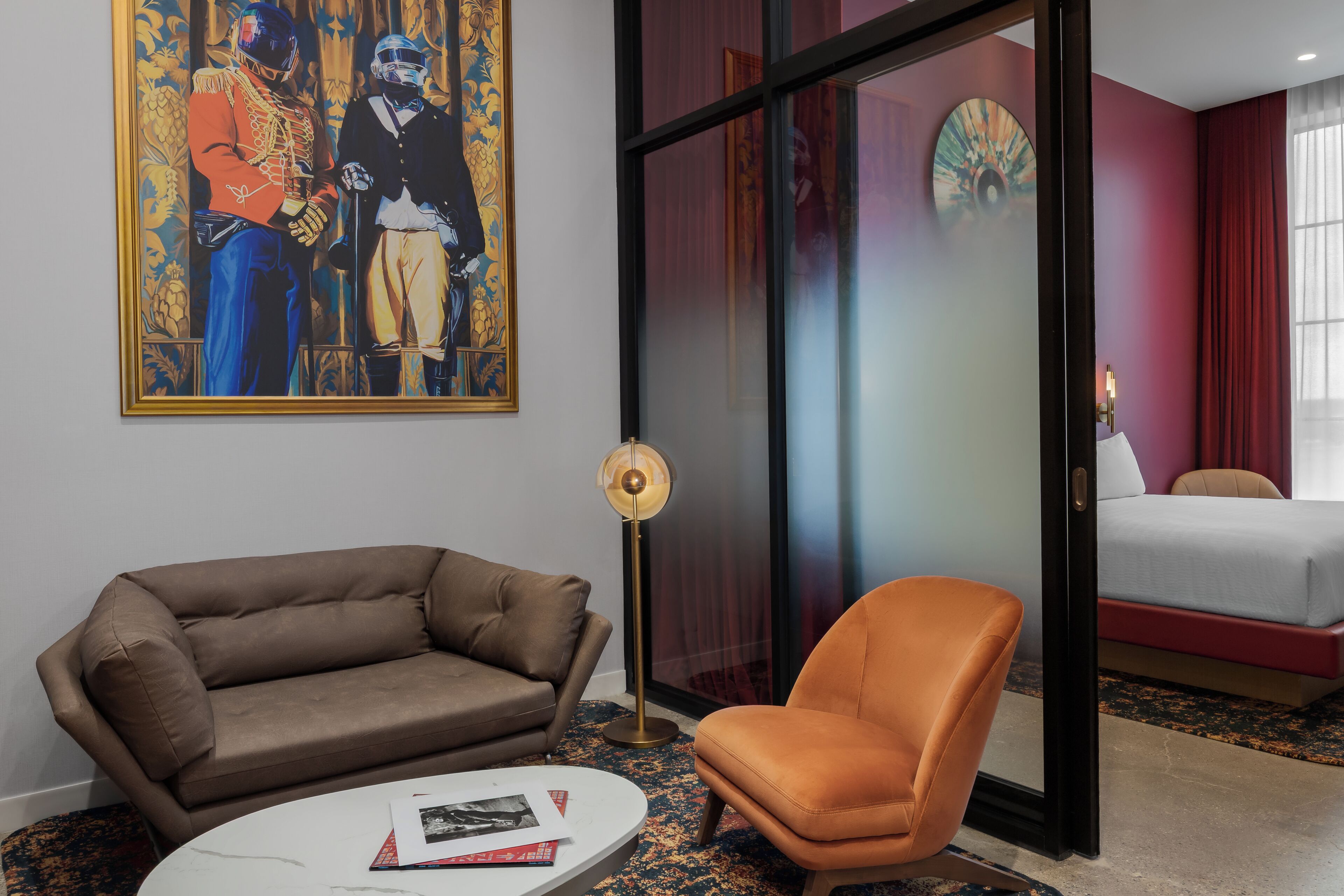 A hotel room with a brown sofa, an orange armchair, a round coffee table, a floor lamp, and a painting on the wall.